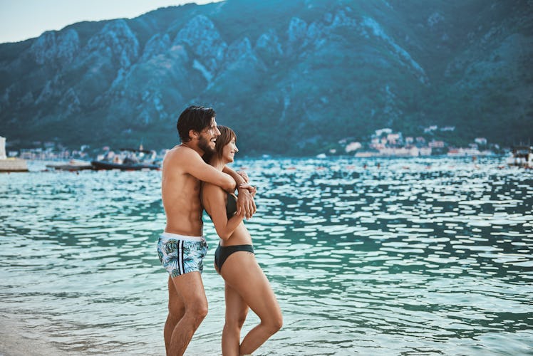A couple embraces while sticking their feet in teal water and looking at blue mountains.