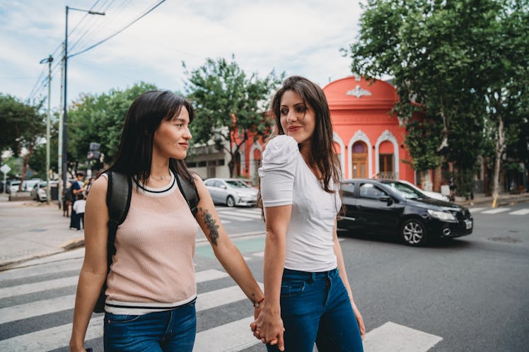 A couple holds hands and walks in a city with a bright coral building in the background.