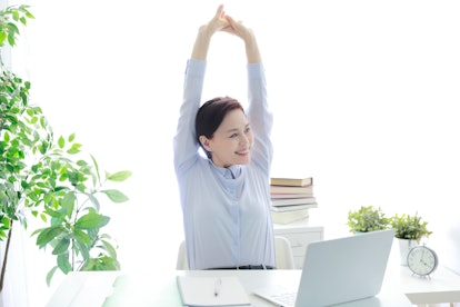 A person sits at a white desk with indoor plants and stretches her arms above her head. Reaching you...