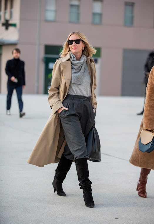 Lisa Aiken in a beige trench coat, grey top and scarf, and black trousers and boots