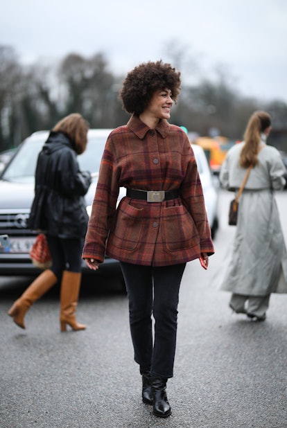 A curly-haired woman in a red-brown checked jacket with belt, black pants and boots at the Copenhage...