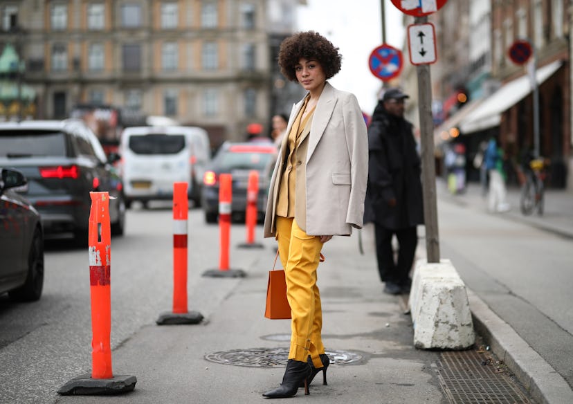 A woman with curly hair in a yellow suit, a beige blazer, black boots and an orange bag at the Copen...
