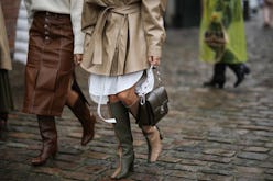 A woman in a beige trench coat, and brown boots and a woman in a brown skirt and white sweater at th...