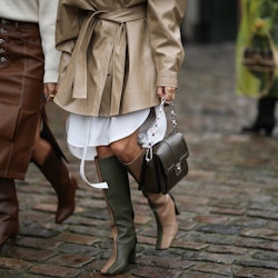 A woman in a beige trench coat, and brown boots and a woman in a brown skirt and white sweater at th...