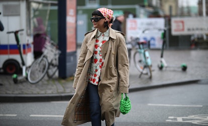 A woman in a white-red top, beige trench coat, red-white headscarf, denim jeans, and a green bag at ...