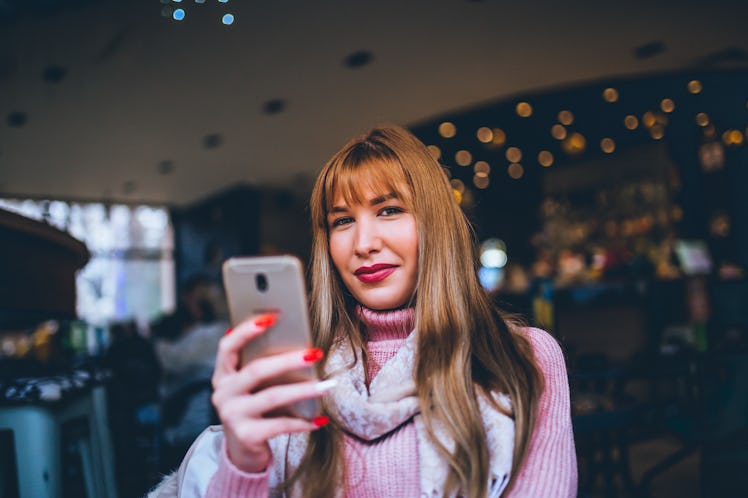 A young woman in a pink turtleneck sits in a coffee shop and holds up her phone.