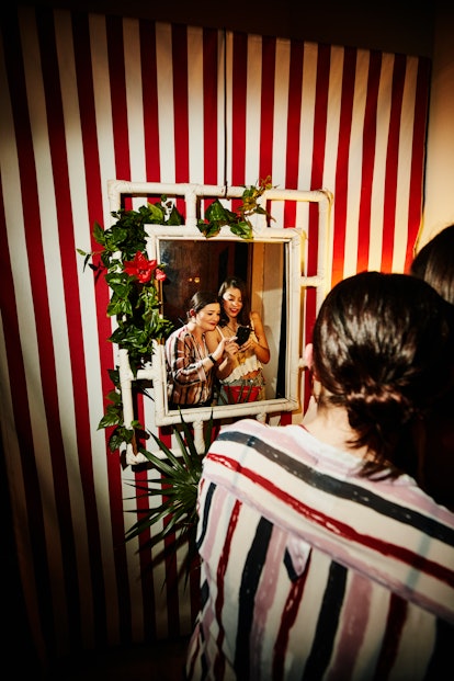 Two women smile and pose in front of a cool mirror on a striped wall for a selfie.