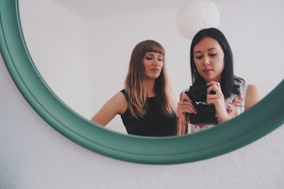 Two women pose for a mirror selfie in a mirror on the wall.