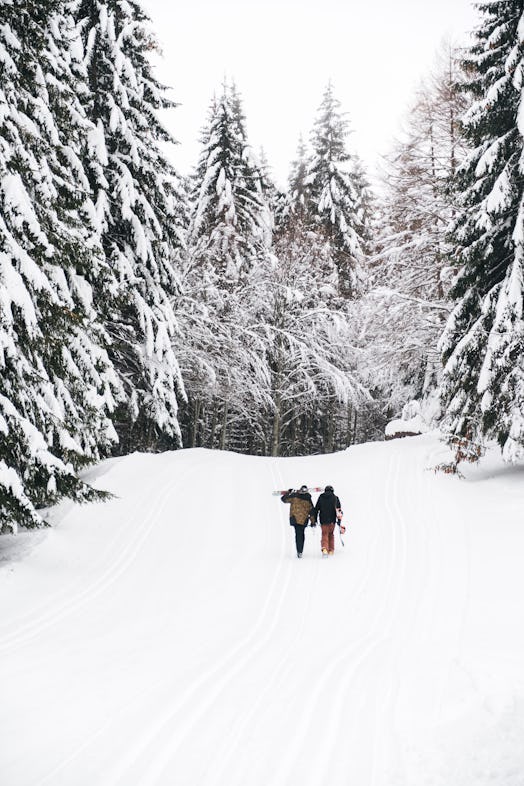 A couple walks with their skis on a snowy path that's lined with evergreen trees.