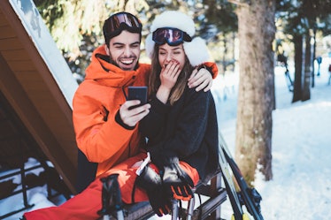 A couple laughs while taking a selfie on their phone during a ski trip.