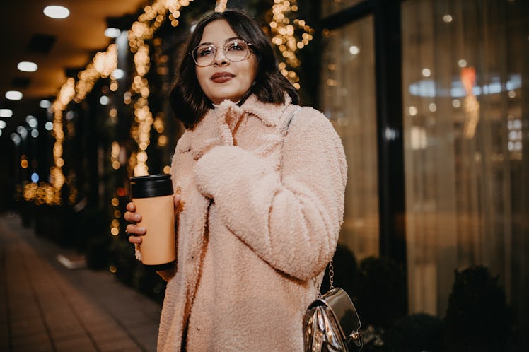 A brunette woman wearing glasses and a light pink coat stands on a sidewalk at night in the city.