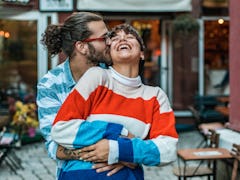 A hipster-looking couple hugs and laughs at an outdoor café.
