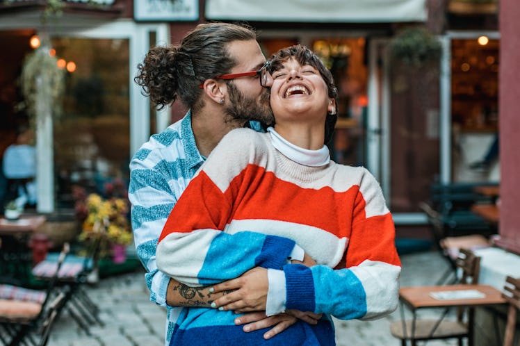 A hipster-looking couple hugs and laughs at an outdoor café.