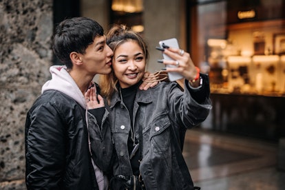 A young, stylish couple takes a flirty selfie on a phone on a rainy day in the city.