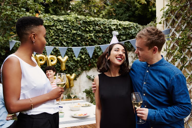 Three friends hang out at a surprise 25th birthday party with glasses of white wine in their hands.
