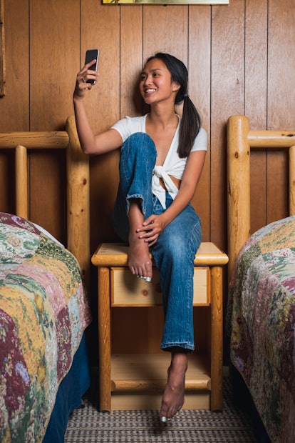 A young Asian woman takes a selfie on her phone while sitting on a wooden nightstand in between two ...