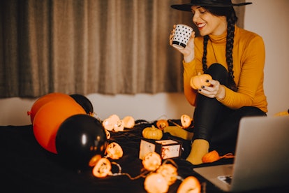 A woman in a witch hat, surrounded by Halloween decorations, holds a cup of coffee. 