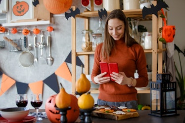 A young woman stands with her phone next to Halloween decorations and cookies set up in her kitchen.