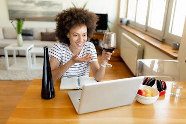 A young Black woman laughs while video chatting on her laptop and drinking a glass of red wine as sh...