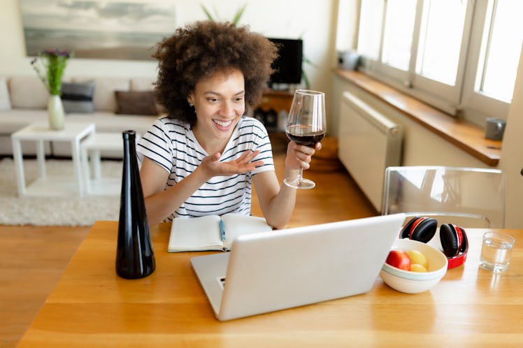 A young Black woman laughs while video chatting on her laptop and drinking a glass of red wine as sh...