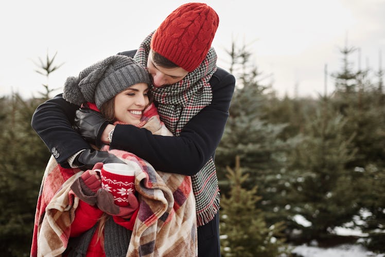 A couple wrapped up in a holiday blanket, embrace outside with a mug of coffee. 