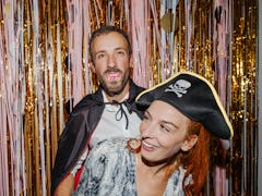 A young couple laughs while wearing Halloween costumes and standing in front of pink and gold stream...