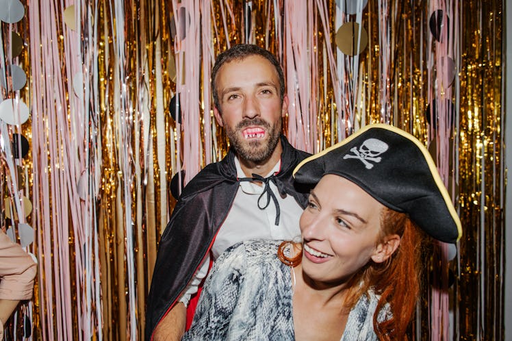 A young couple laughs while wearing Halloween costumes and standing in front of pink and gold stream...