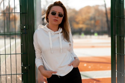 A young stylish woman poses in a white hoodie in front of a basketball court.