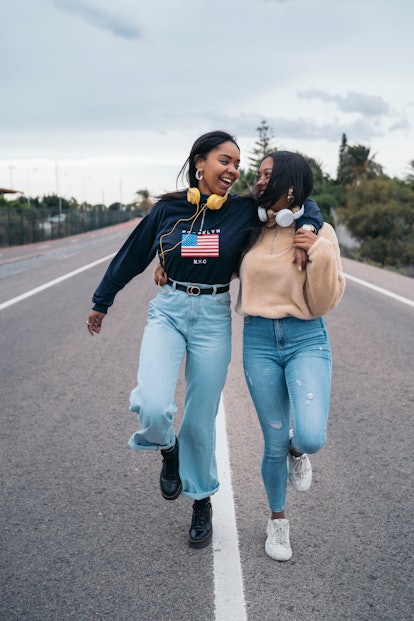 Two young Black women skip on a road while wearing sweatshirts and headphones.