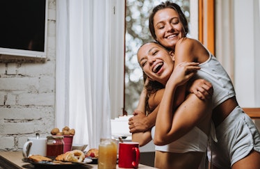 Two young women hug in their kitchen while enjoying breakfast.
