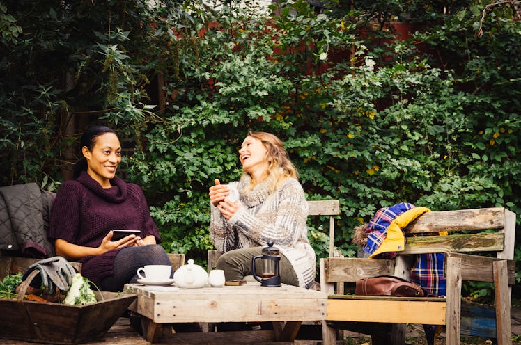 Two young women sit in their backyard and drink tea while wearing sweaters.