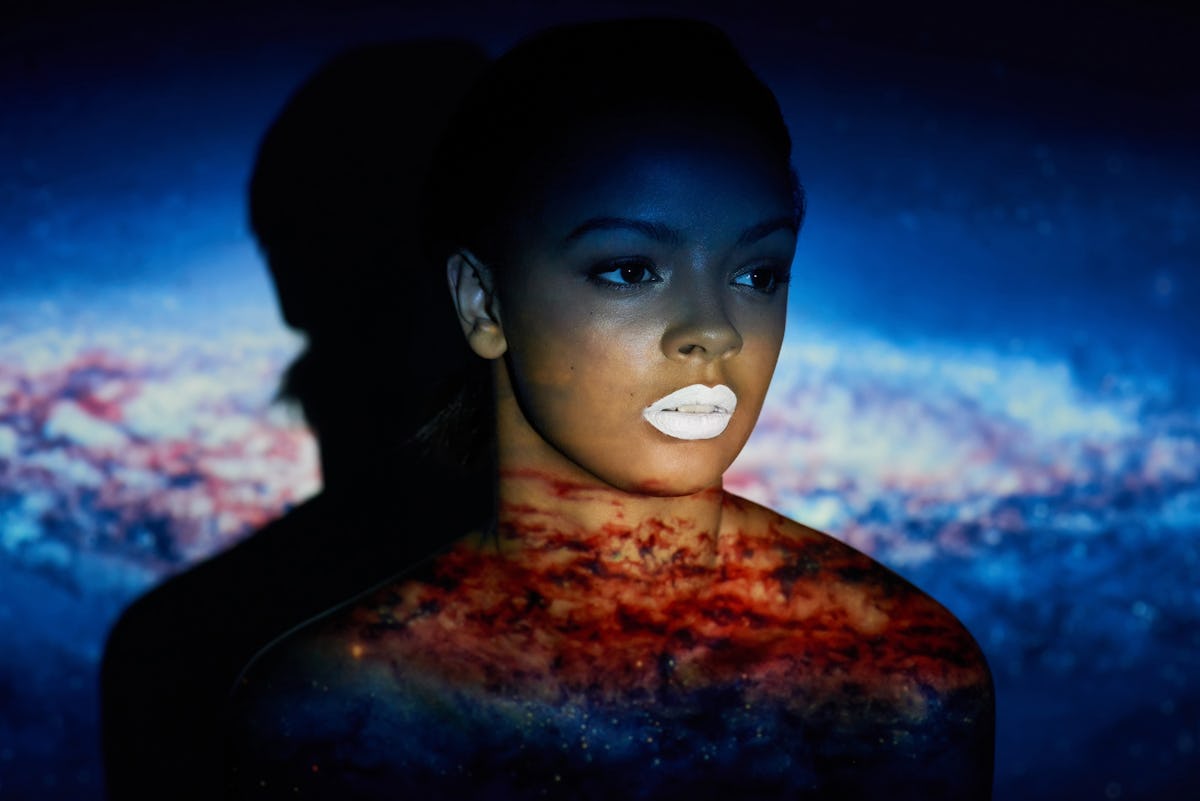 A woman with white lipstick stands in front of a blue and red celestial backdrop.