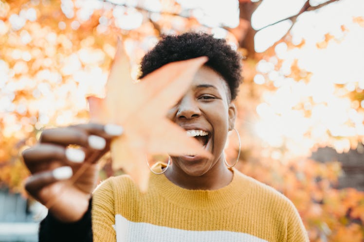 A young Black woman holds up a fall leaf while wearing a sweater and hanging out in a park.