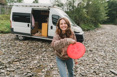 A young woman tosses a frisbee while wearing a sweater and hanging outside her campervan.