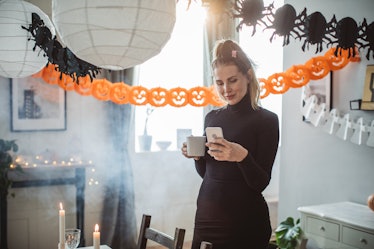 A woman in a black dress holds a cup of coffee and texts while standing in a living room that's deco...