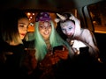 Three friends dressed up for Halloween sit in the backseat of a car while looking at one of their ce...