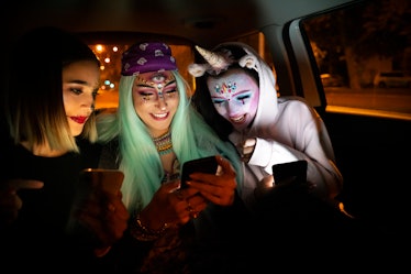 Three friends dressed up for Halloween sit in the backseat of a car while looking at one of their ce...