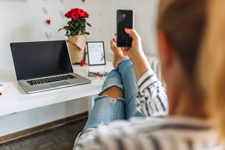 A young woman puts her feet up on her desk while video chatting on her phone next to a bouquet of ro...