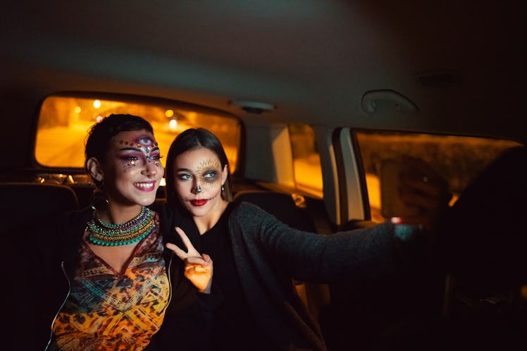Two young women dressed in Halloween costumes sit in the back of their car and take a selfie.
