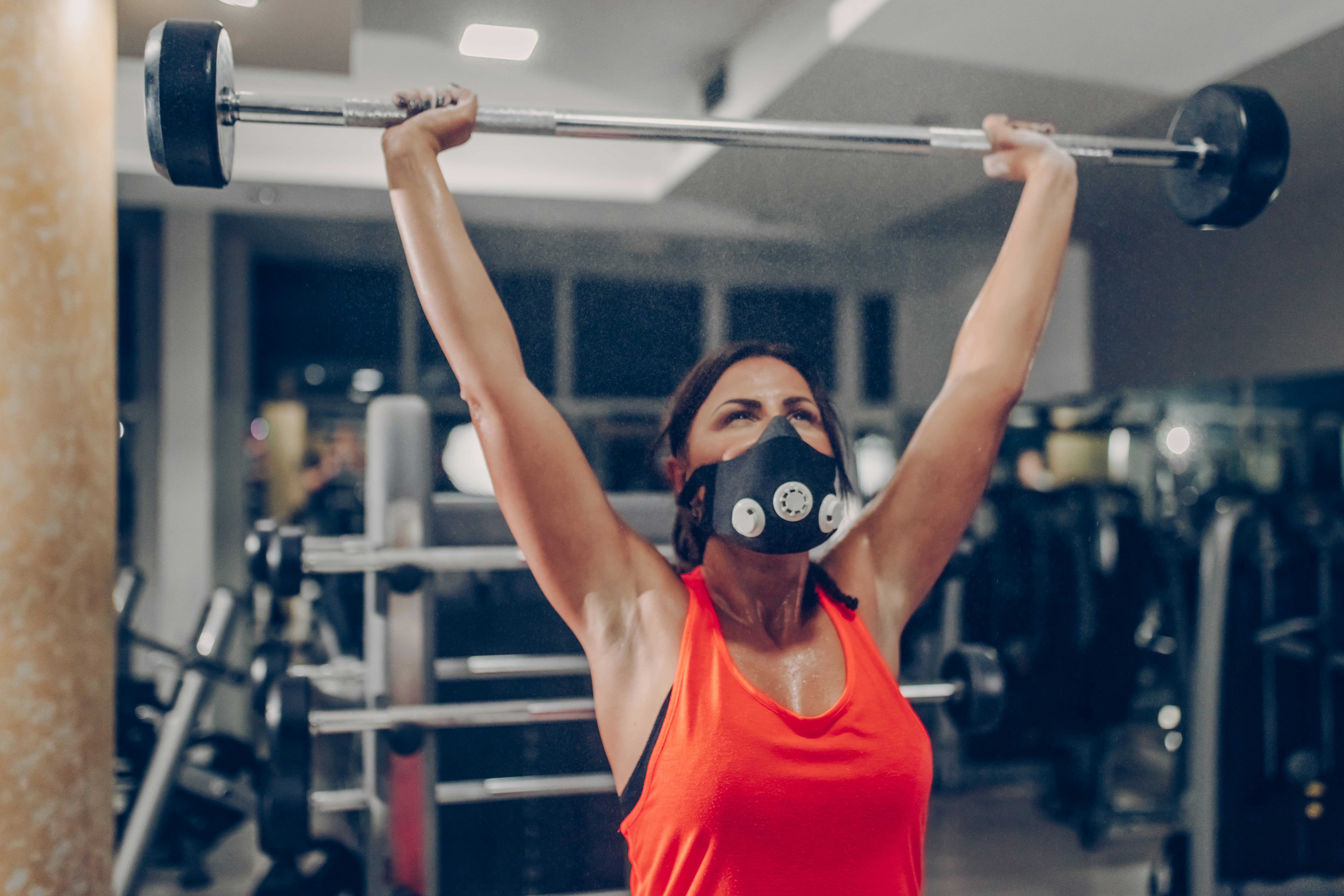 A person wearing an orange tank top and a black mask presses a barbell overhead. Resistance training...