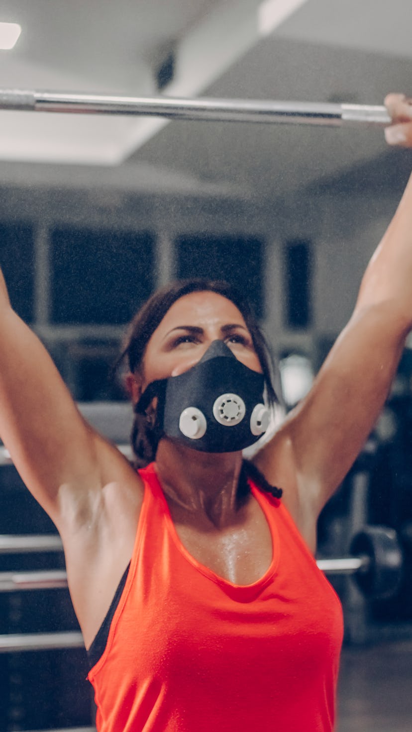 A person wearing an orange tank top and a black mask presses a barbell overhead. Resistance training...