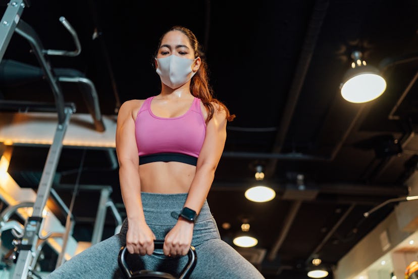 A person wearing a pink sports bra and a mask performs a kettlebell deadlift in the gym. Lifting wei...