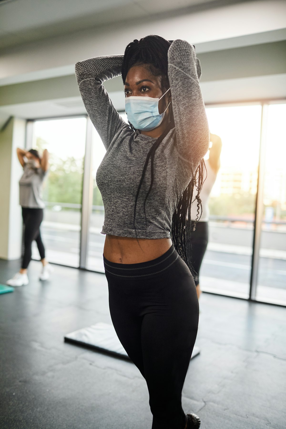 A person wearing a mask and long-sleeved grey shirt performs an overhead tricep extension in a group...