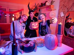 Three friends dressed in Halloween costumes pose for a phone selfie with their jack-o-lantern at hom...