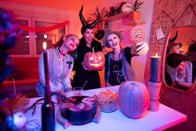 Three friends dressed in Halloween costumes pose for a phone selfie with their jack-o-lantern at hom...
