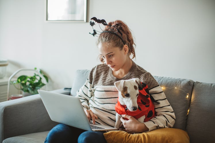 A young woman sits with her dog and her laptop on a couch, while wearing a headband with bats on her...