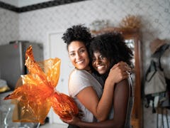 Two friends hug in their kitchen, while one holds an orange wrapped gift. 