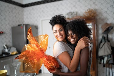 Two friends hug in their kitchen, while one holds an orange wrapped gift.