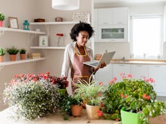 A young Black woman looks at her laptop while standing next to a table filled with potted plants in ...