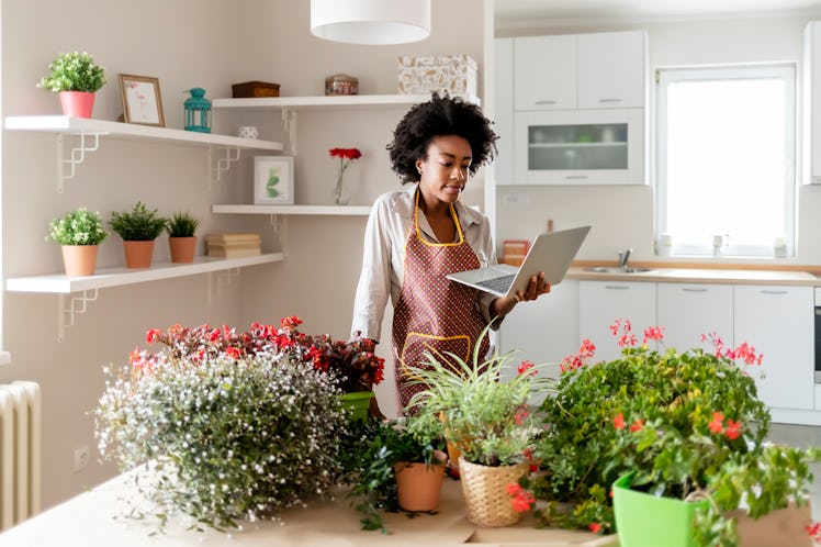 A young Black woman looks at her laptop while standing next to a table filled with potted plants in ...